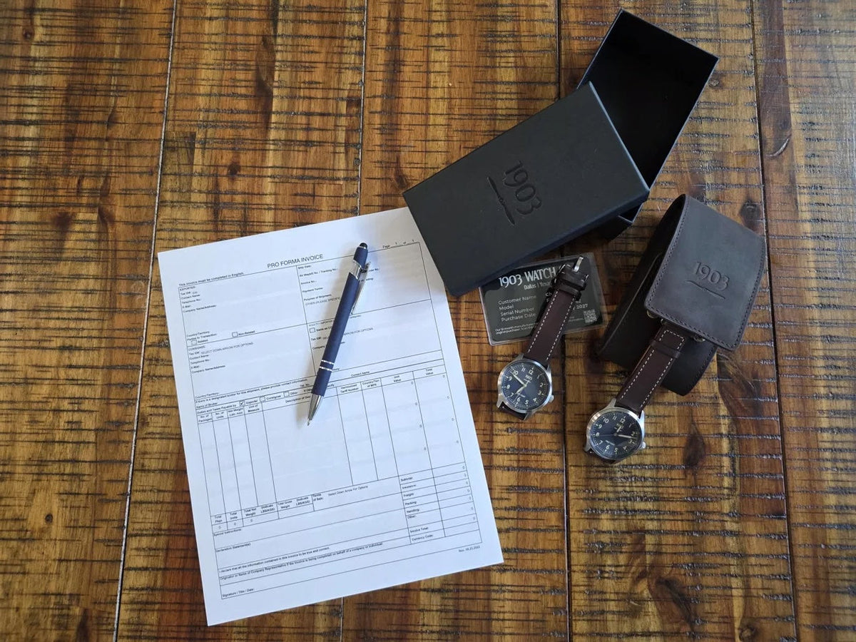Two 1903 Watch Co. First Flight watches, branded packaging, and a customs pro forma invoice on a wood table